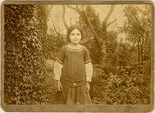 Fotografía antigua en papel tono sepia de una niña posando en un jardín rodeada de vegetación