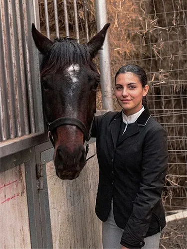 Mujer vestida de amazona junto a un caballo en un picadero o cuadra.