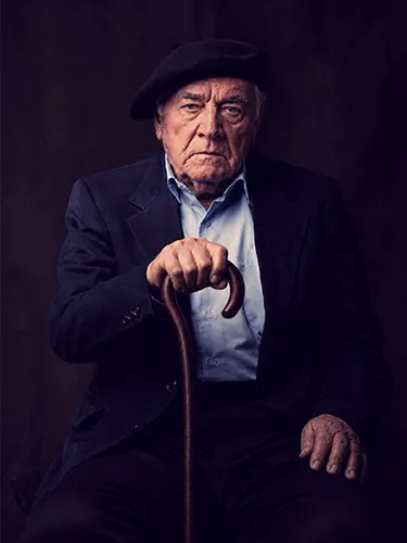 Retrato de un hombre anciano con boina negra y chaqueta, apoyado en un bastón de madera.