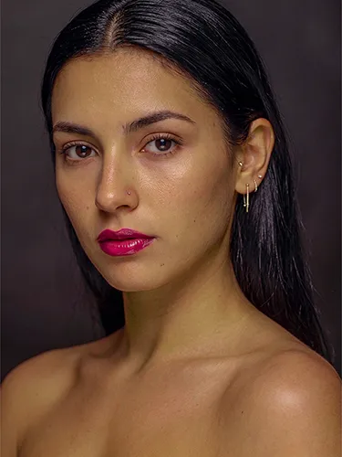 Retrato de estudio de una mujer con pendientes dorados, piercing en la nariz y labios fucsia.