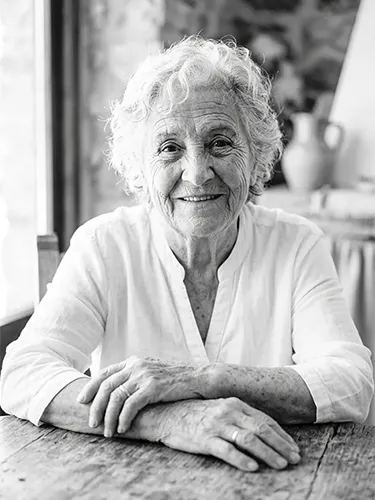 Retrato en blanco y negro de una mujer mayor sonriente sentada ante una mesa de madera.