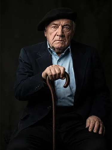 Retrato artístico con iluminación dramática de un anciano con traje oscuro, boina y bastón de madera.